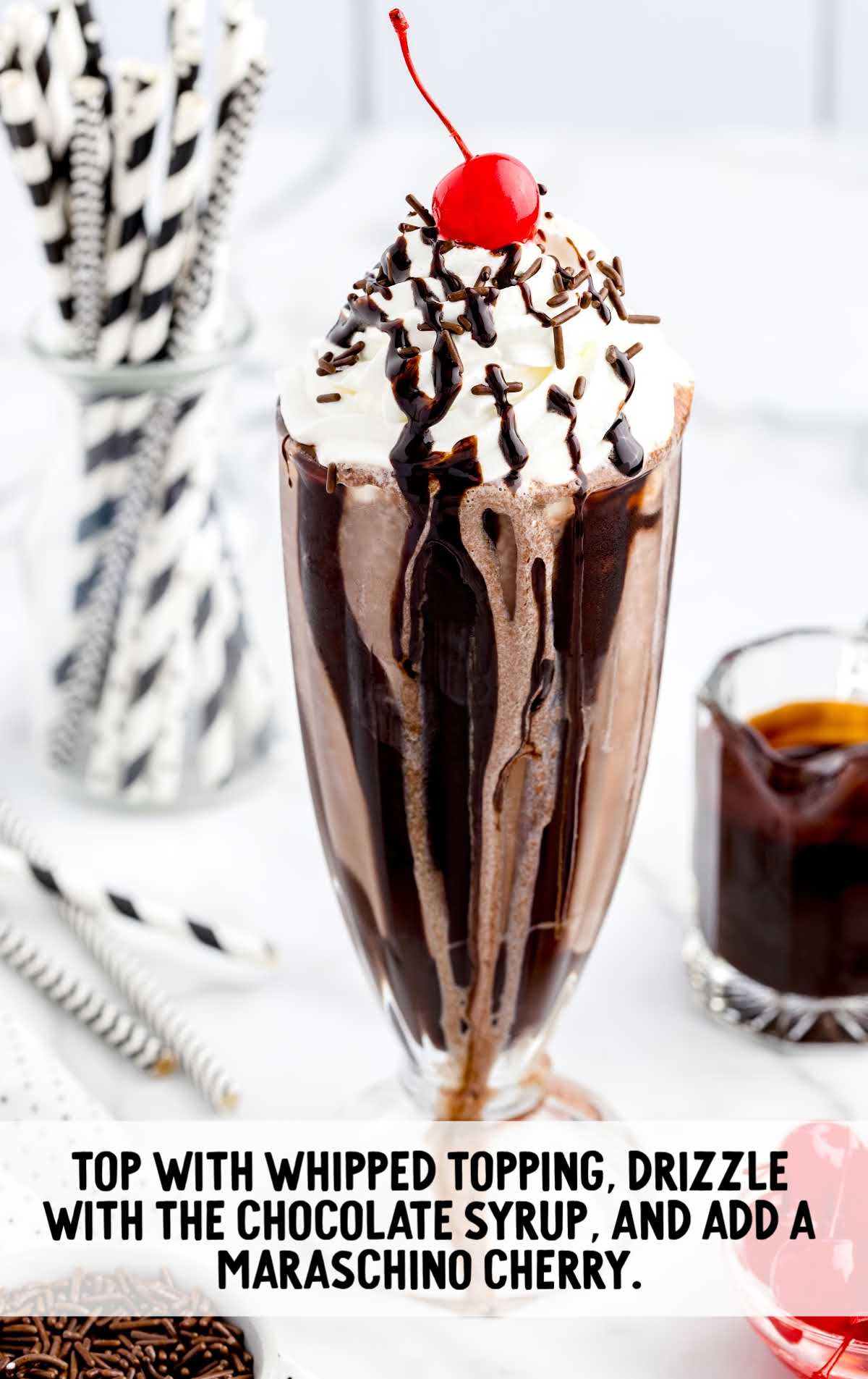  a tall glass topped with whipped cream then garnished with Hershey bars, a cherry, chocolate sprinkles, and chocolate syrup