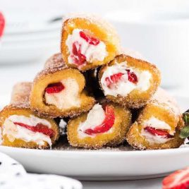 close up shot of French Toast Roll-Ups rolled in cinnamon sugar piled on a plate with slices of strawberries