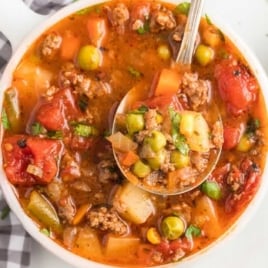 Bowl of hearty hamburger soup filled with ground beef, potatoes, peas, corn, and tomatoes with a spoon lifting a serving.