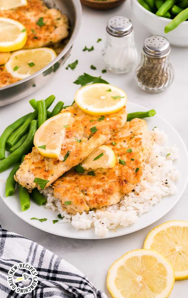 close up shot of Lemon Chicken with lemon slices and garnished with parsley served with a side of white rice and green beans on a plate