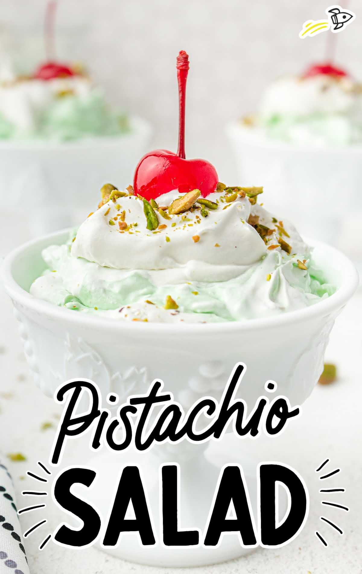 close up shot of a small bowl of pistachio salad topped with whipped cream and crushed pistachios with a cherry on top