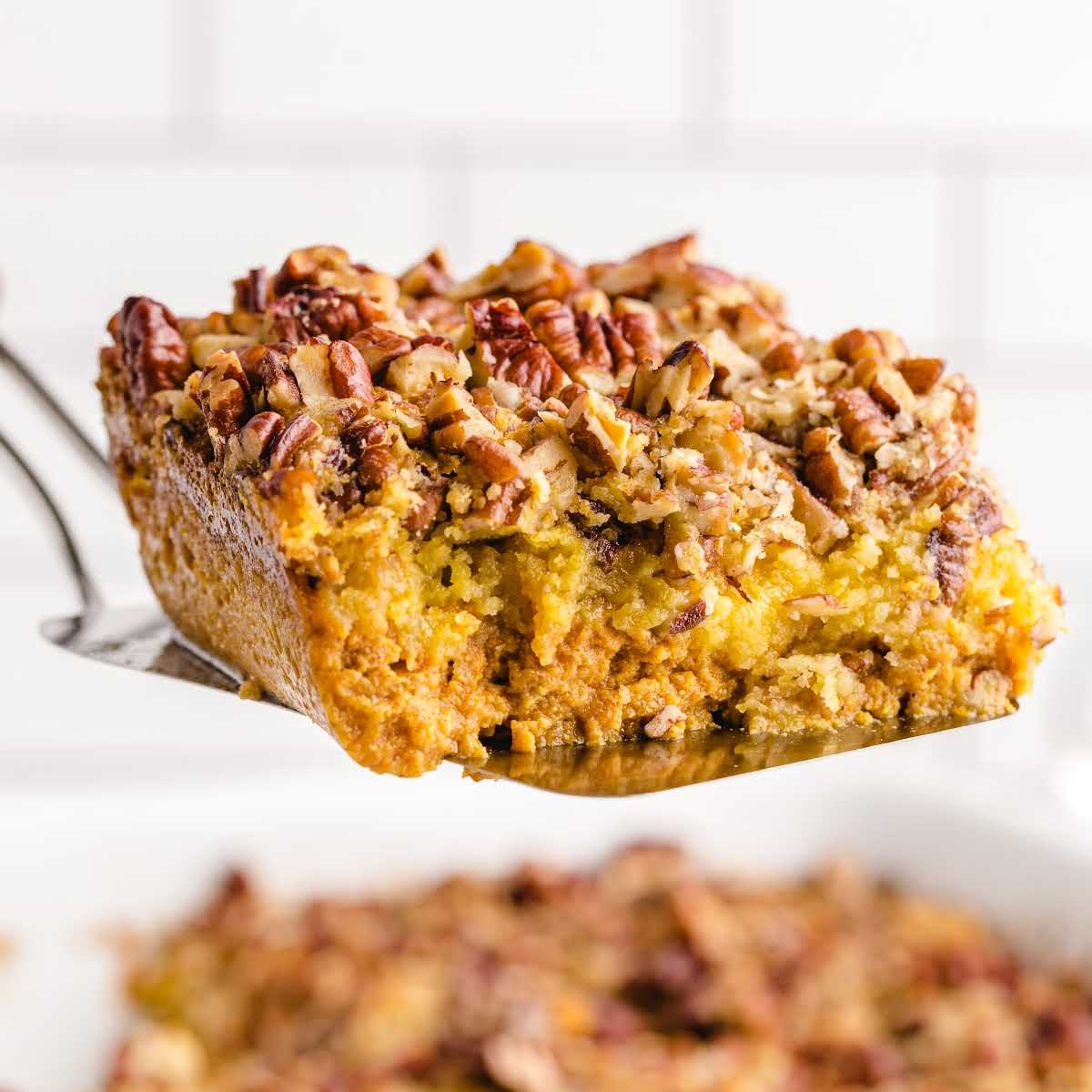 Close-up of a slice of pumpkin crunch cake being lifted from a baking dish showing moist pumpkin base and crunchy pecan topping