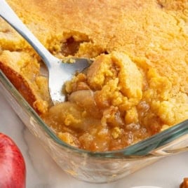 A spoon scoops out a serving of golden brown Caramel Apple Dump Cake from a glass baking dish, showing the baked crust and caramelized apple filling inside.