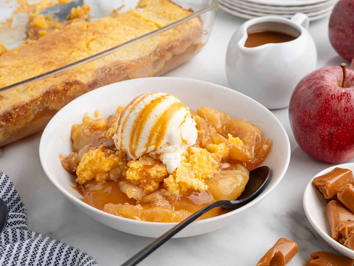 A bowl of apple cobbler topped with vanilla ice cream and caramel sauce sits on a table, reminiscent of a classic Caramel Apple Dump Cake. In the background are a spoon, apples, caramel candies, and a baking dish of cobbler, evoking the comforting flavors found in any delicious Caramel Apple Dump Cake Recipe.