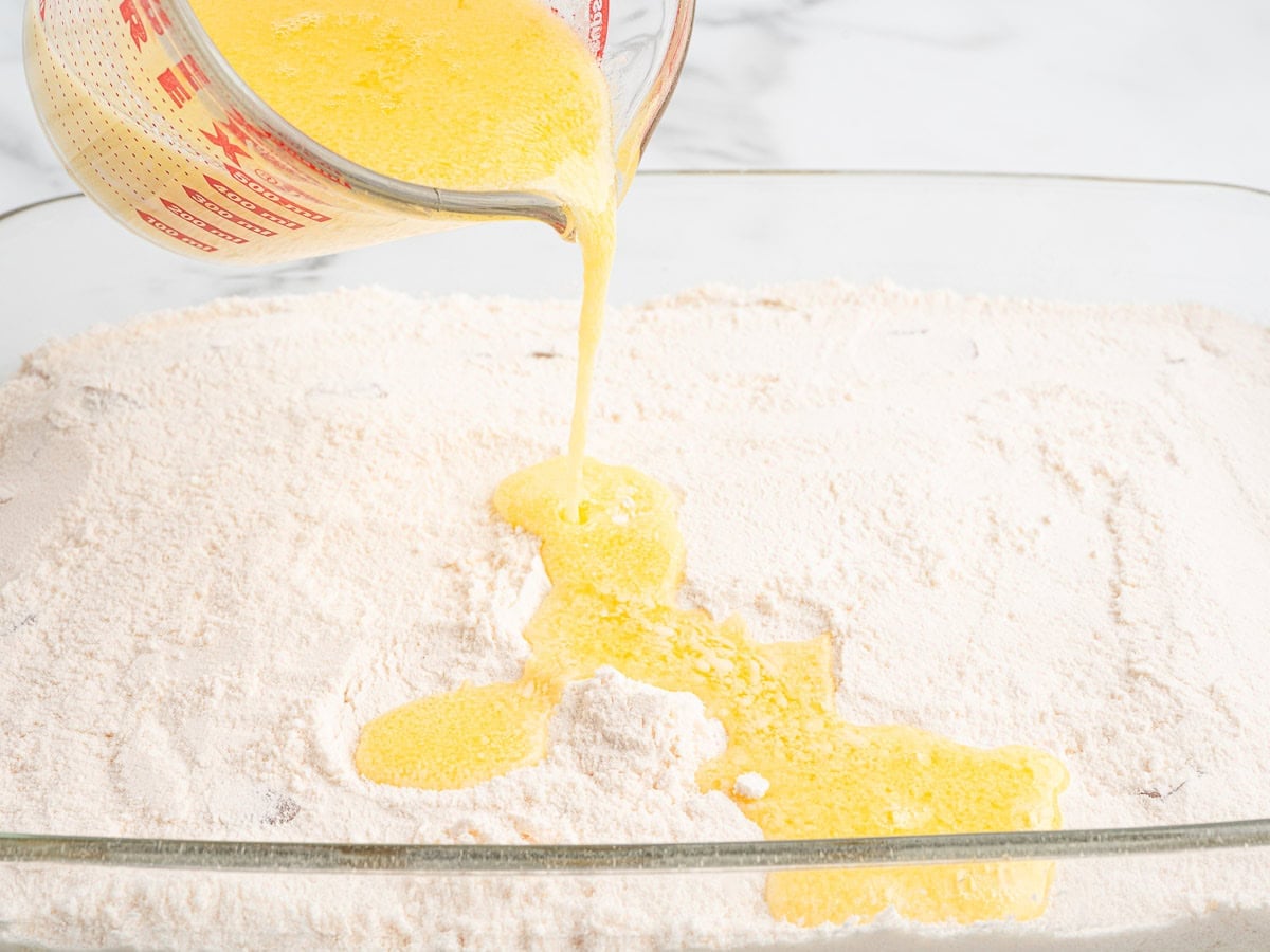 Melted butter is poured from a measuring cup onto a layer of dry cake mix in a glass baking dish on a white countertop, preparing the base for a delicious Caramel Apple Dump Cake.