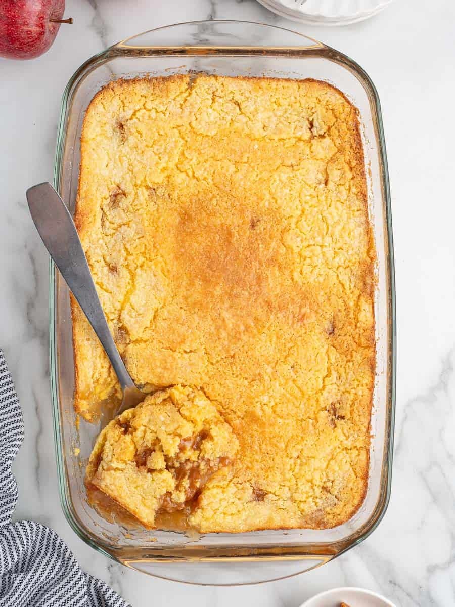 A glass baking dish filled with golden-brown Caramel Apple Dump Cake, one corner scooped out with a metal serving spoon, revealing a gooey, caramel-like filling inside. The dish sits on a marble surface with a striped cloth nearby.