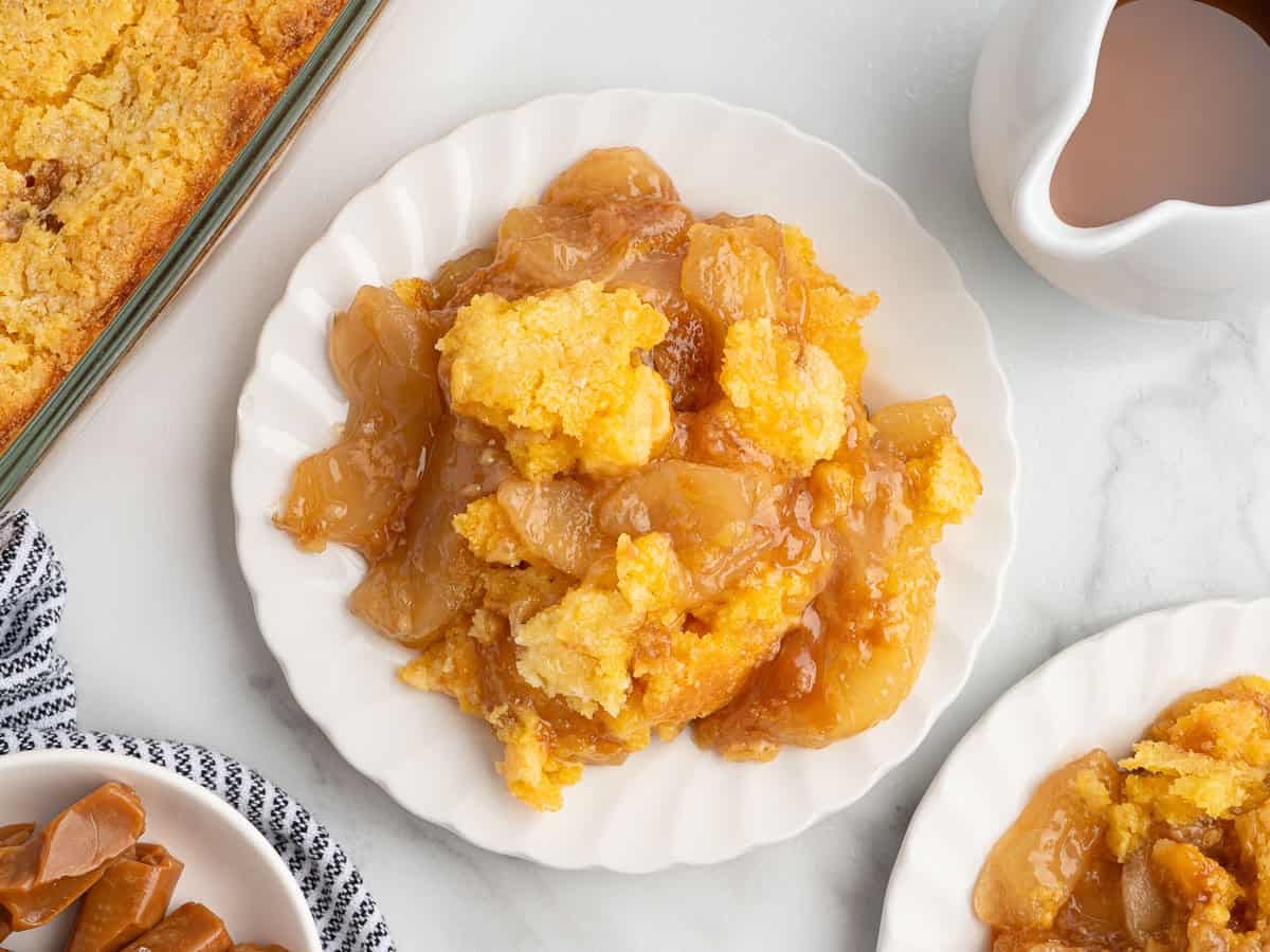 A serving of apple cobbler with a golden crumb topping is on a white plate, surrounded by more cobbler, caramel candies, and a small pitcher of sauce on a light surface—perfect for anyone craving the cozy flavors of a Caramel Apple Dump Cake.