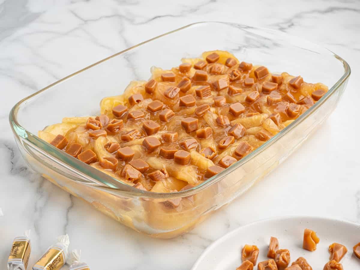 A glass baking dish filled with sliced apples topped with small caramel candy pieces, ready to be baked, sits on a white marble countertop—perfect for making a delicious Caramel Apple Dump Cake Recipe.