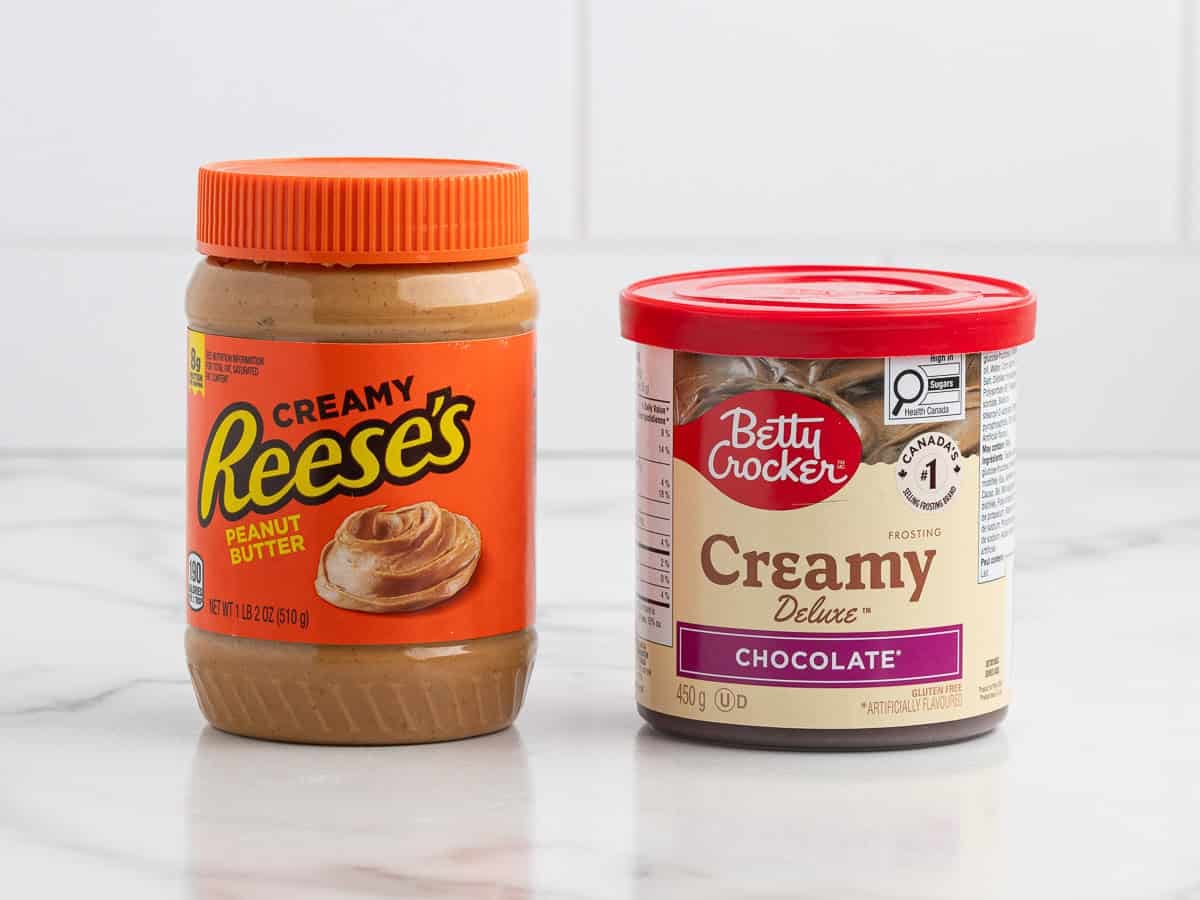 A jar of Reeses creamy peanut butter and a tub of Betty Crocker creamy chocolate frosting sit side by side on a white marble countertop with a white tiled background, ready to be transformed into a delicious Chocolate Peanut Butter Fudge.