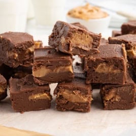A stack of chocolate fudge squares with a visible peanut butter swirl sits on parchment paper, offering a tempting preview of this irresistible Chocolate Peanut Butter Fudge Recipe, while glasses of milk and a bowl of peanut butter add to the inviting scene in the background.