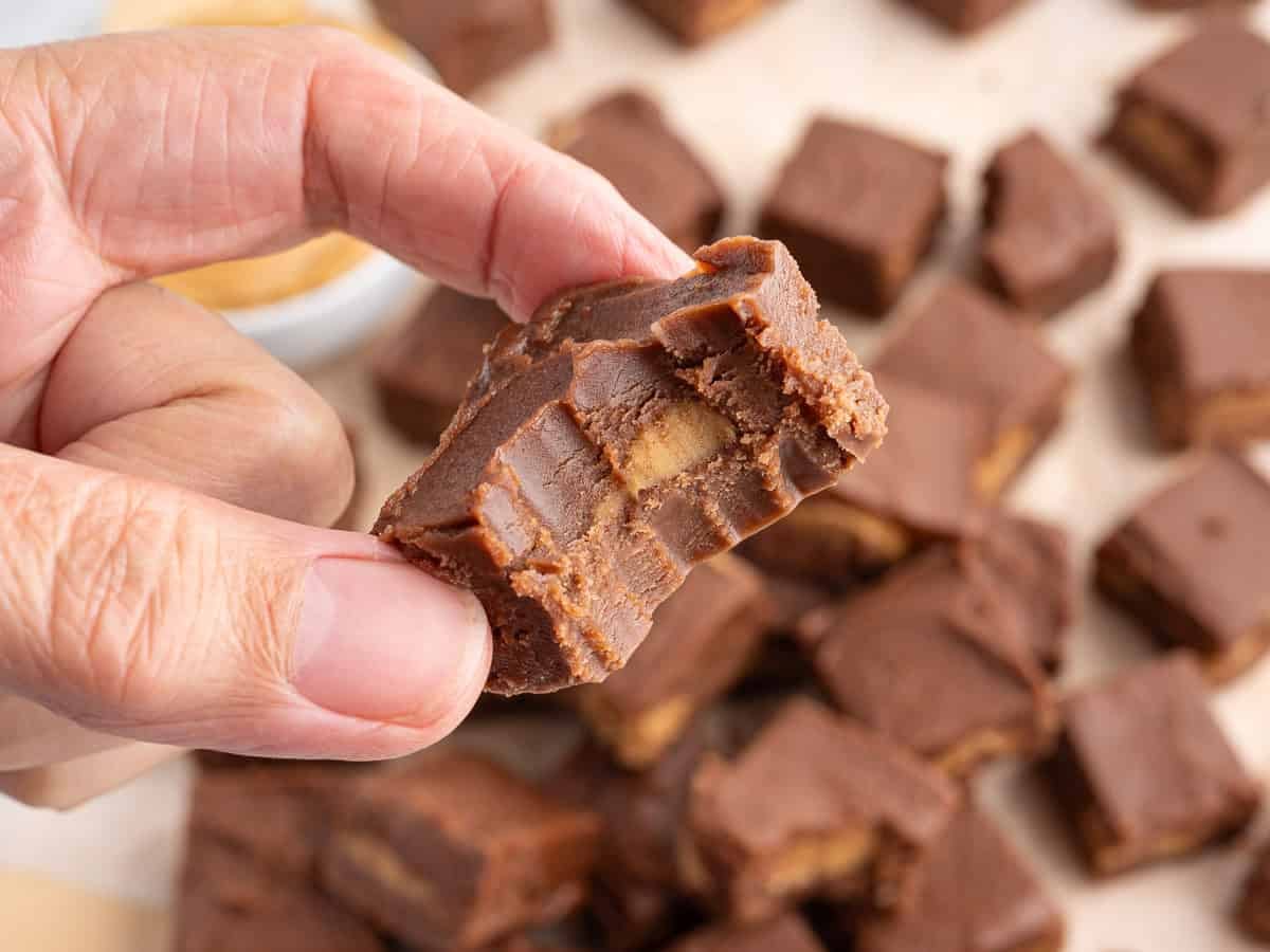 A hand holding a piece of chocolate fudge with a bite taken out, revealing a creamy peanut butter filling—this irresistible treat is the result of our signature Chocolate Peanut Butter Fudge Recipe. More pieces of the same decadent Chocolate Peanut Butter Fudge are blurred in the background.