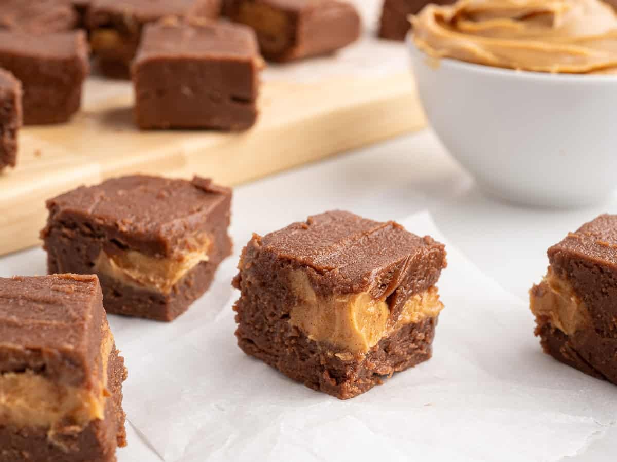 Squares of chocolate and peanut butter fudge sit on parchment, with more Chocolate Peanut Butter Fudge and a bowl of peanut butter in the background—making this scene irresistible for anyone searching for an easy Chocolate Peanut Butter Fudge recipe.
