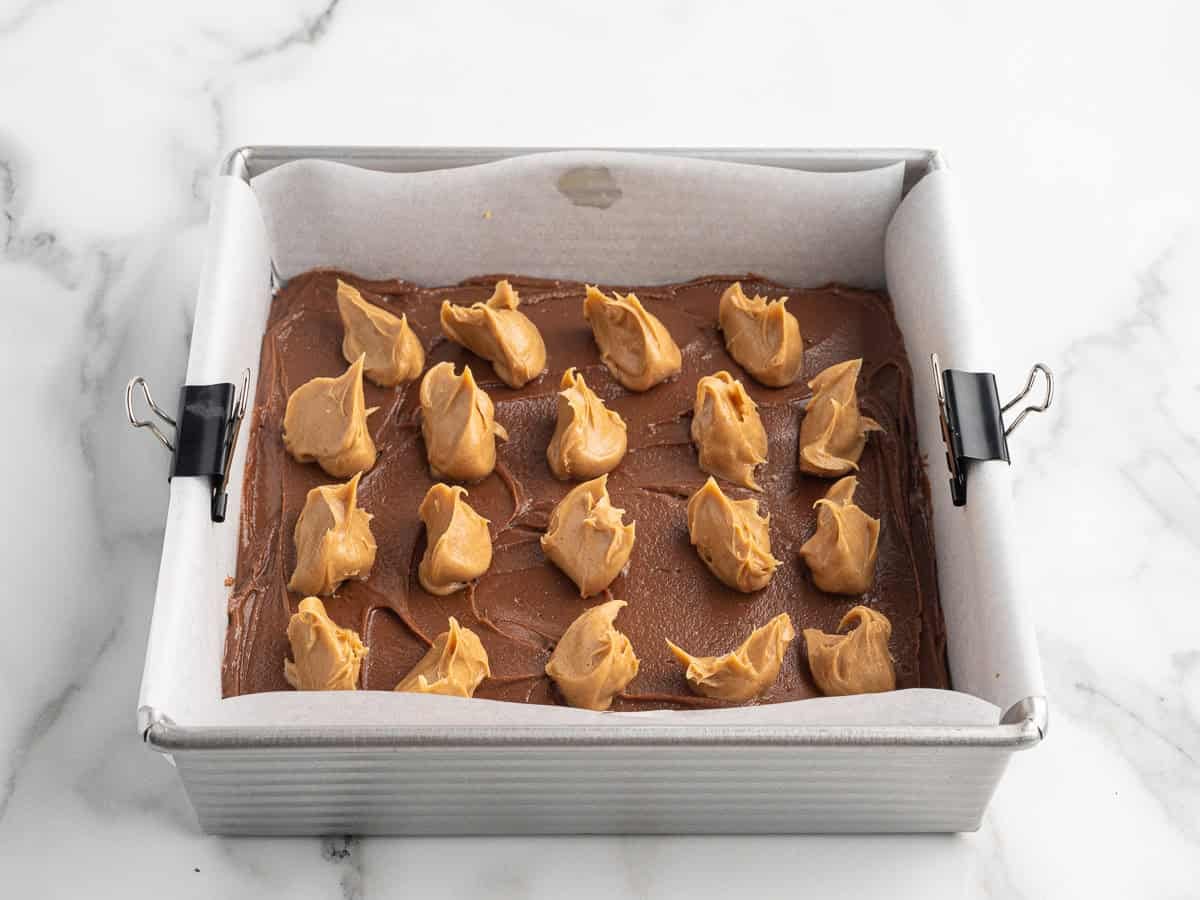 A square baking pan lined with parchment paper holds rich chocolate batter topped with dollops of creamy peanut butter, ready to be swirled for the perfect Chocolate Peanut Butter Fudge. Black binder clips secure the parchment to the pan.