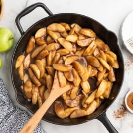 Sliced apples cooked in a skillet with cinnamon sauce, being stirred with a wooden spoon—this classic Fried Apples Recipe fills the air with warmth and spice. Green apples, a gray towel, and a small bowl of cinnamon are nearby on a white countertop.