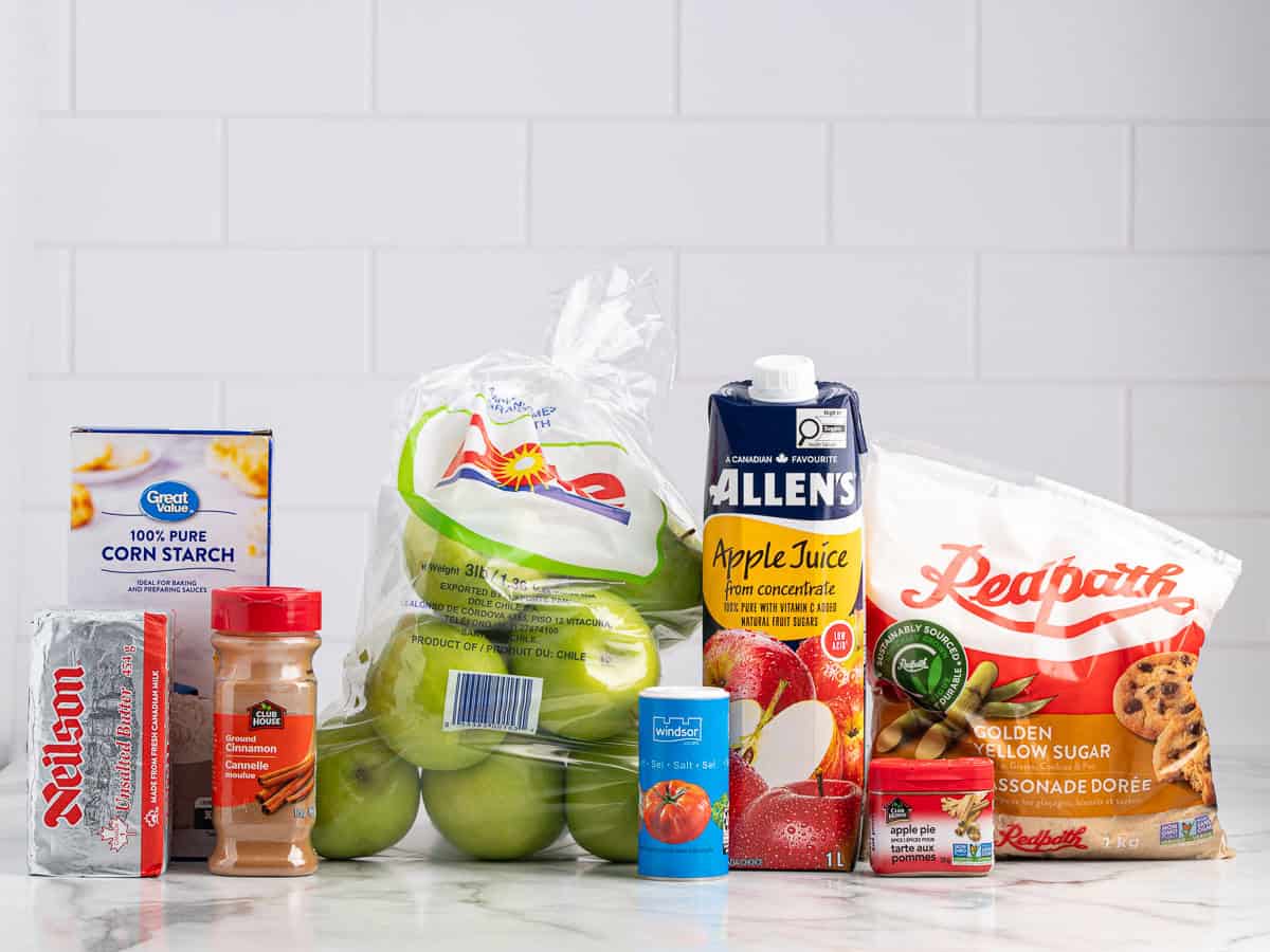 A variety of grocery items on a counter, including green apples, apple juice, butter, cinnamon, corn starch, brown sugar, and canned milk—everything you need to make a classic Fried Apples Recipe—arranged against a white tiled background.