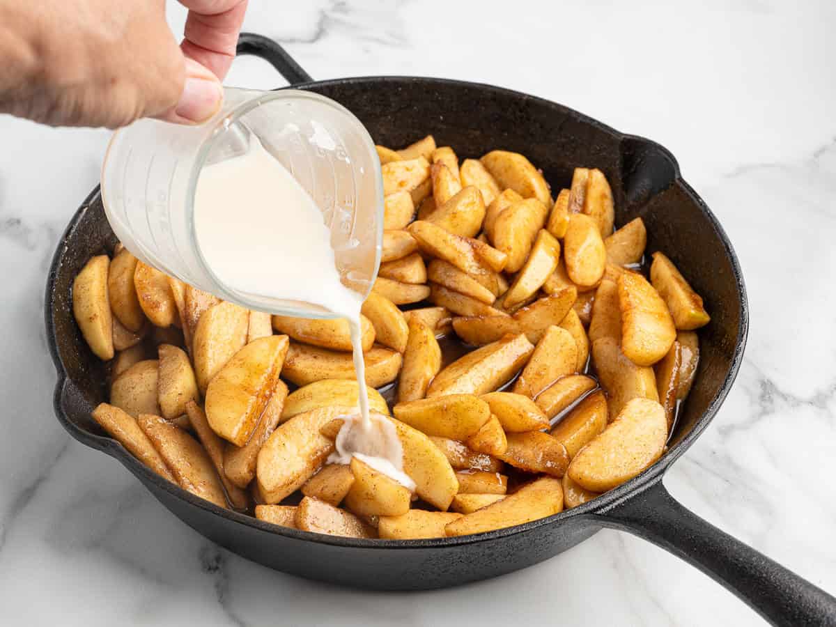 A hand pours a creamy liquid from a measuring cup onto sliced, cooked apples in a black cast iron skillet on a marble surface, creating the perfect finishing touch for this classic Fried Apples Recipe.