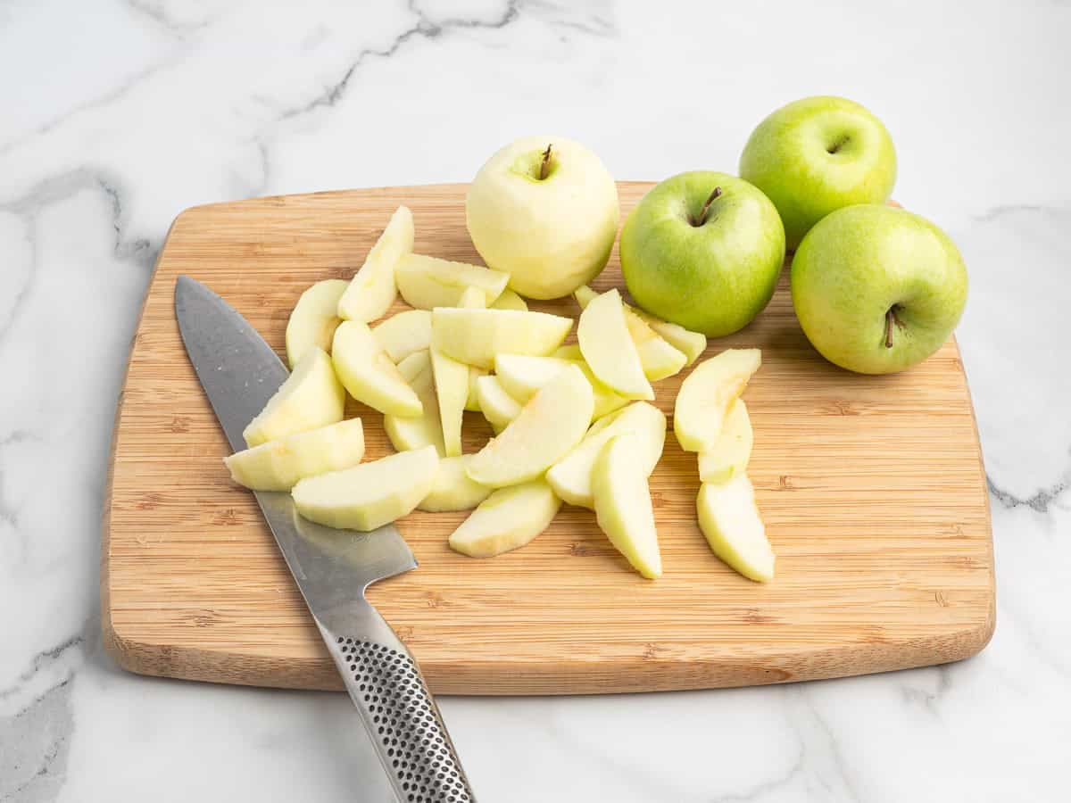 A knife and sliced green apples on a wooden cutting board, with whole green apples and a peeled apple nearby, are arranged on a marble surface—perfect for preparing your favorite Fried Apples recipe.