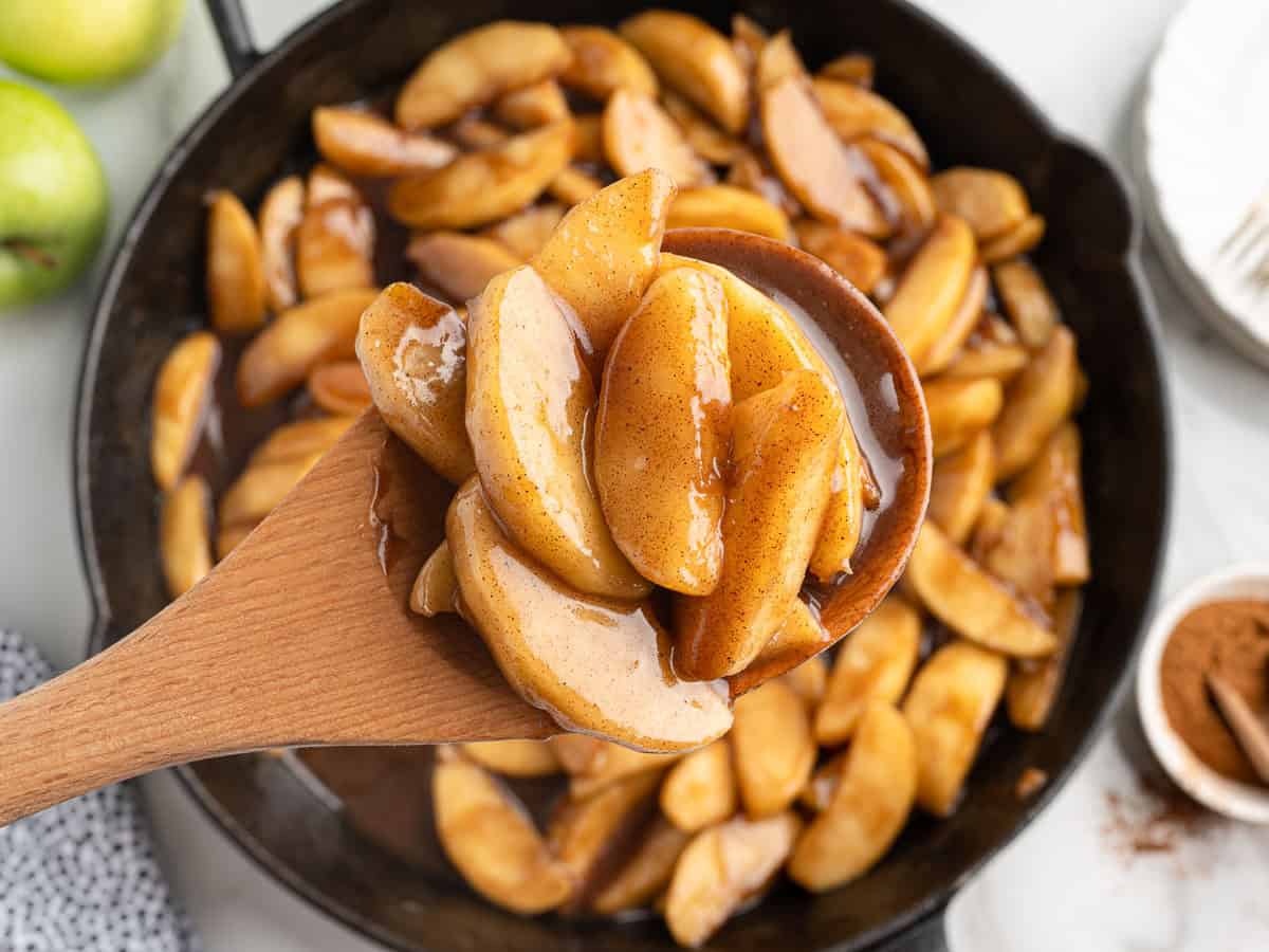 A wooden spoon holds cooked apple slices with cinnamon above a skillet filled with more apple slices, showcasing this delicious Fried Apples Recipe. A bowl of cinnamon and whole green apples are visible in the background.