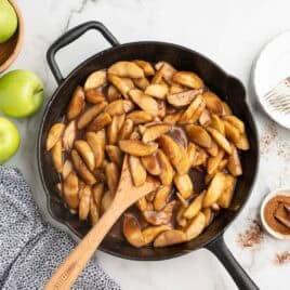 A cast iron skillet filled with cooked cinnamon apples sits on a marble surface, showcasing a classic fried apples recipe. Nearby are green apples, a bowl of cinnamon, a plate with forks, and a blue patterned cloth. A wooden spoon rests in the skillet.
