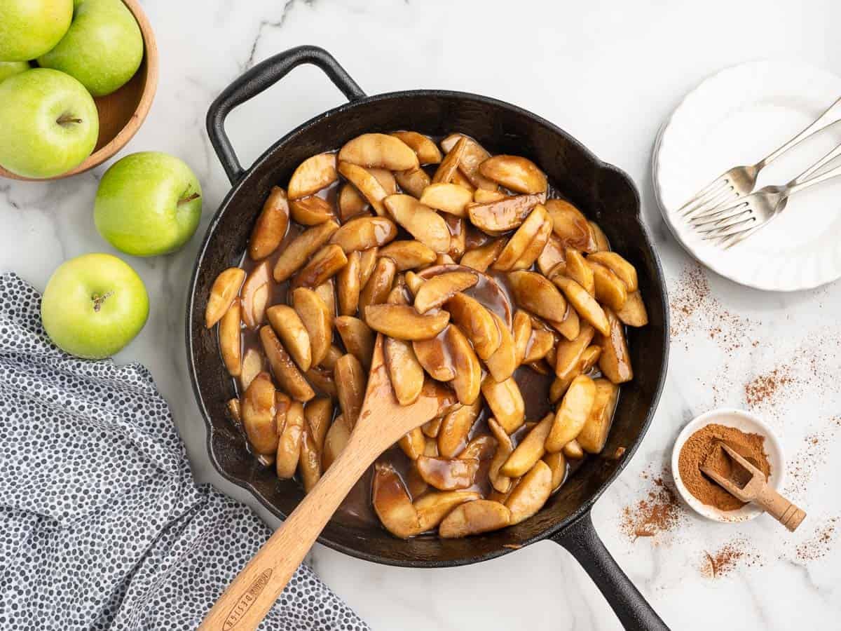 A cast iron skillet filled with cooked cinnamon apples sits on a marble surface, showcasing a classic fried apples recipe. Nearby are green apples, a bowl of cinnamon, a plate with forks, and a blue patterned cloth. A wooden spoon rests in the skillet.