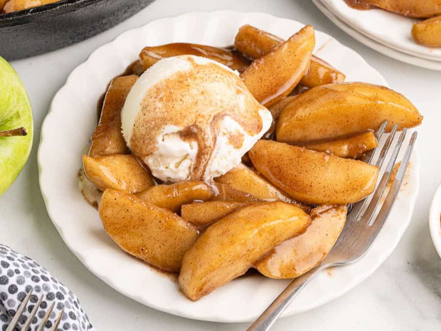 A white plate with sliced baked apples inspired by a classic Fried Apples Recipe, covered in cinnamon sauce and topped with a scoop of vanilla ice cream. A fork rests on the plate.