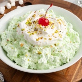 A bowl of light green Pistachio Salad topped with whipped cream, chopped pistachios, and a maraschino cherry sits on a wooden surface, with mini marshmallows nearby&mdash;an inviting Pistachio Salad Recipe for any gathering.