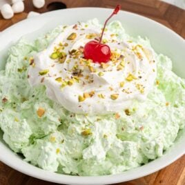A bowl of green Pistachio Salad topped with whipped cream, chopped pistachios, and a red cherry sits on a wooden surface, mini marshmallows scattered beside it—perfect for anyone craving a classic Pistachio Salad Recipe.