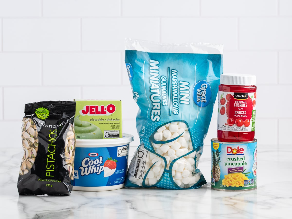 A delightful Pistachio Salad Recipe with ingredients displayed on a white surface: Cool Whip, pistachio Jell-O pudding mix, pistachios, mini marshmallows, crushed pineapple, and maraschino cherries.