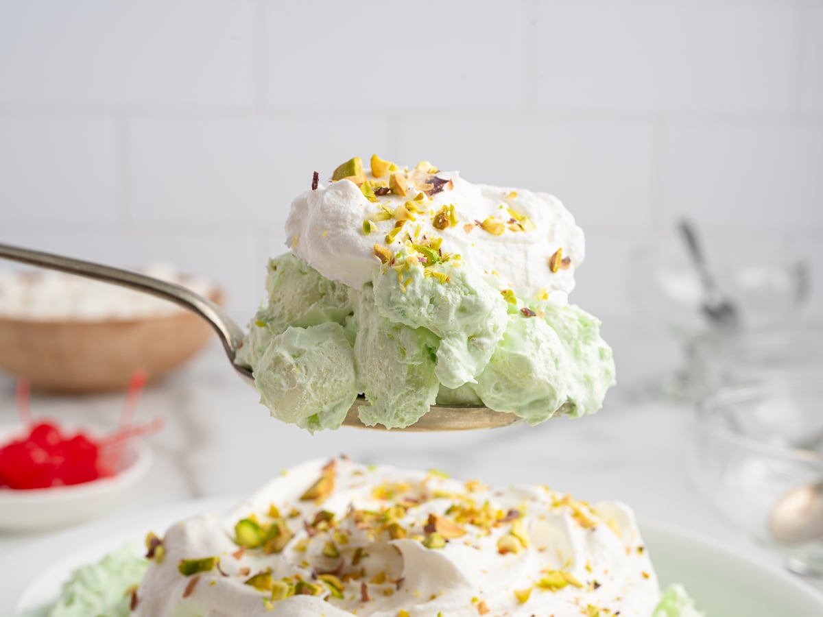 A large spoon holds a scoop of green Pistachio Salad topped with whipped cream and chopped pistachios, showcasing this classic Pistachio Salad Recipe, with a plateful and blurred cherries in the background.