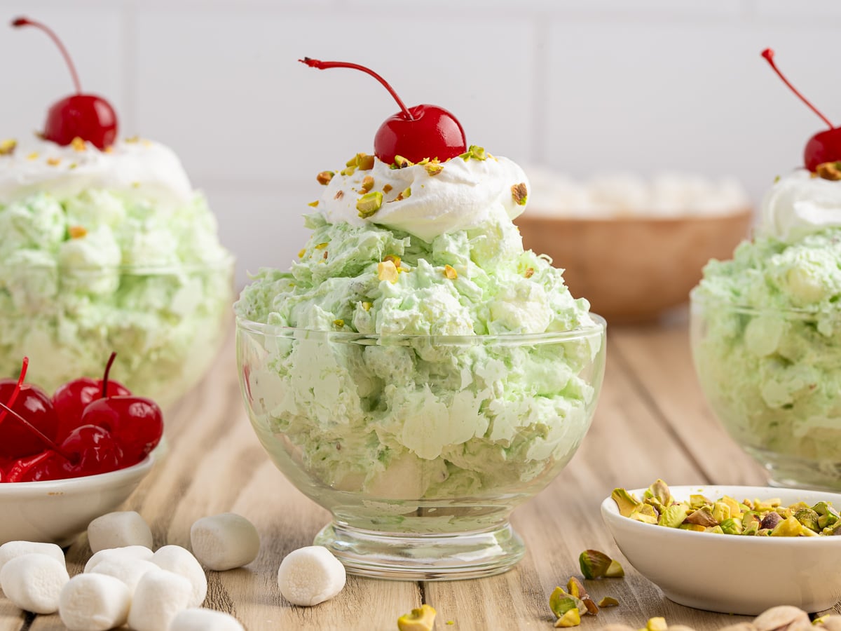 A glass dish filled with green Pistachio Salad, topped with whipped cream, chopped pistachios, and a maraschino cherry. Surrounding bowls hold ingredients for this classic Pistachio Salad Recipe—marshmallows, cherries, and pistachios on a wooden surface.