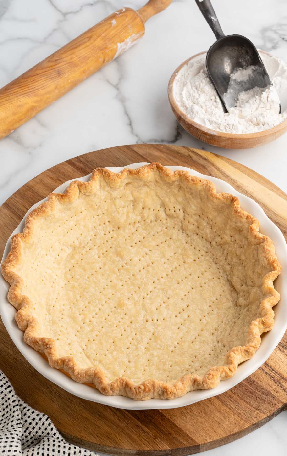 Golden baked pie crust with fork pricks in the bottom, rolling pin and bowl of flour in the background