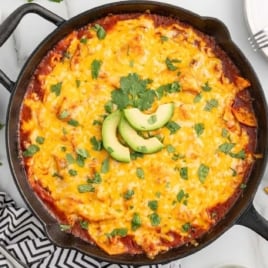 Overhead view of a skillet filled with baked chicken enchiladas, topped with melted cheese, avocado slices, and fresh cilantro.