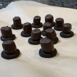 Twelve chocolate-covered marshmallows are placed on top of chocolate cookie halves, arranged on a beige surface, resembling charming Snowmen Truffles ready for your next Snowmen Truffles Recipe.