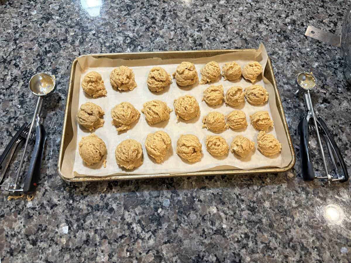 A baking sheet with twenty dough balls, perfect for a batch of Snowmen Truffles, rests on parchment paper atop a granite countertop, with two ice cream scoops placed on either side of the sheet.