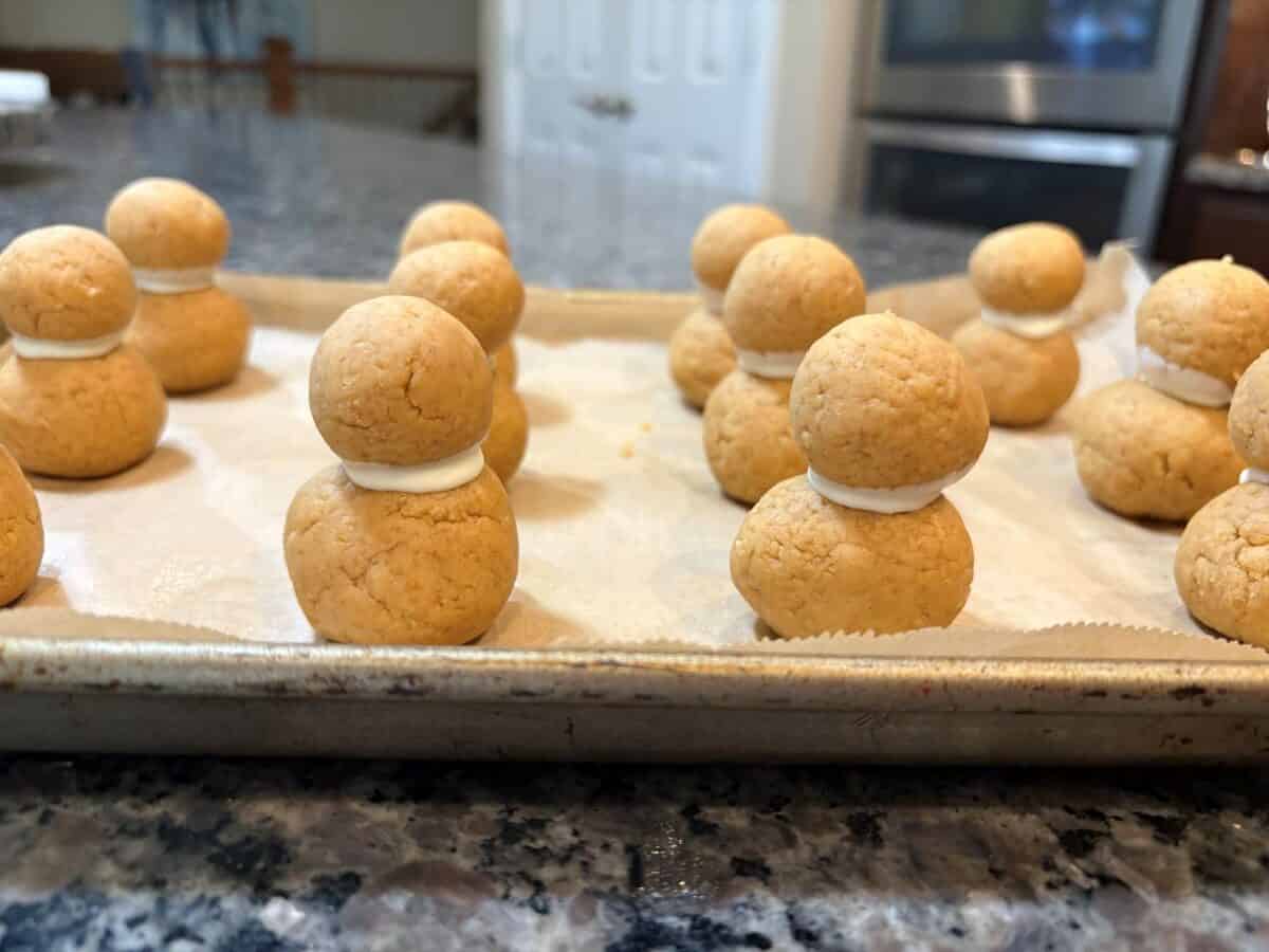 Round golden cookies stacked in pairs with white filling between, arranged on a parchment-lined baking sheet on a kitchen counter—reminiscent of the playful spirit found in a Snowmen Truffles Recipe. Ovens and cabinets are visible in the background.