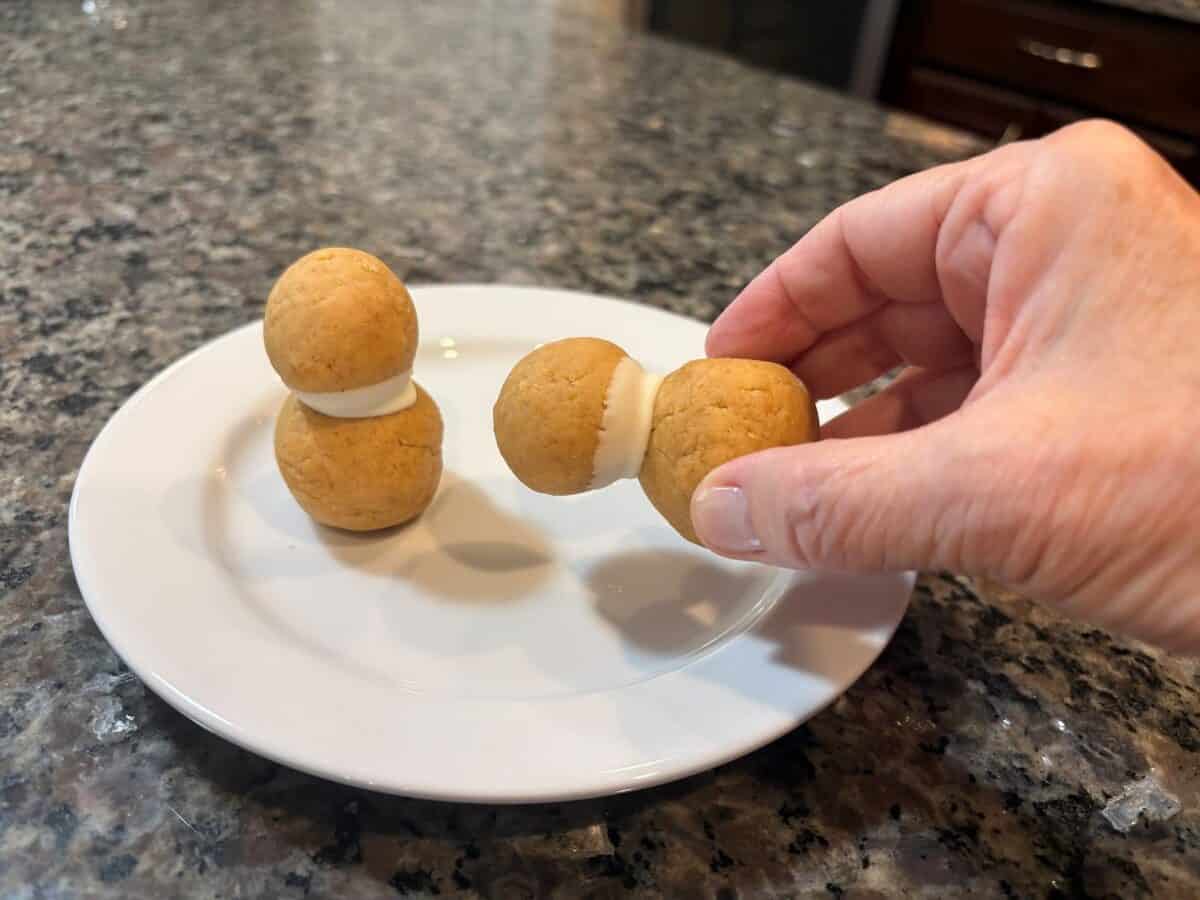A hand holds a peanut butter ball sandwich with white filling, reminiscent of Snowmen Truffles, next to another similar treat on a white plate on a granite countertop.