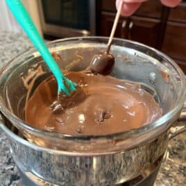 A hand dips a cherry with a toothpick into melted chocolate in a double boiler on a kitchen counter, just like the first step in creating delicious Snowmen Truffles. A spatula rests in the chocolate, ready to help with this delightful Snowmen Truffles Recipe.