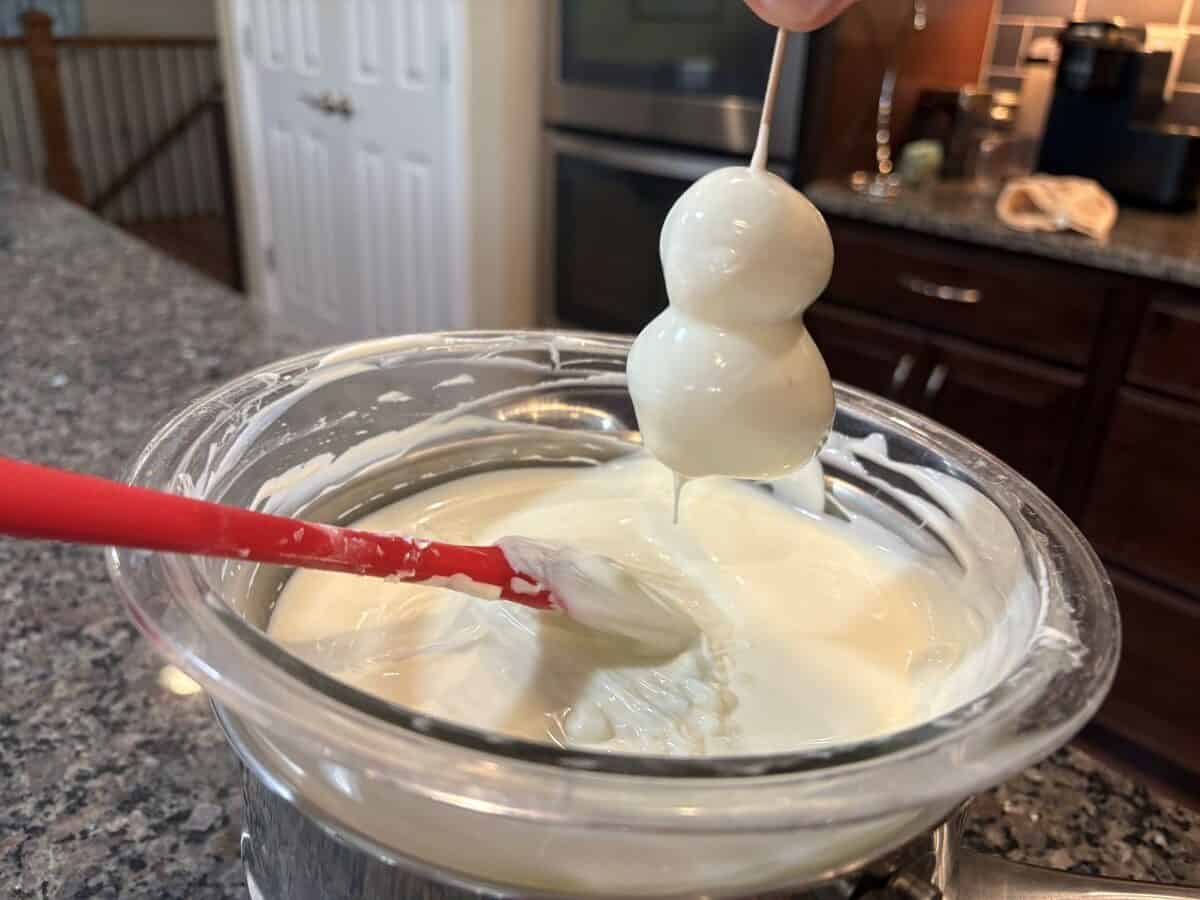 A hand holds a stick with two round treats dipped in white chocolate over a bowl of melted white chocolate; a red spatula rests in the bowl on a kitchen counter, ready for the next step of this delightful Snowmen Truffles Recipe.