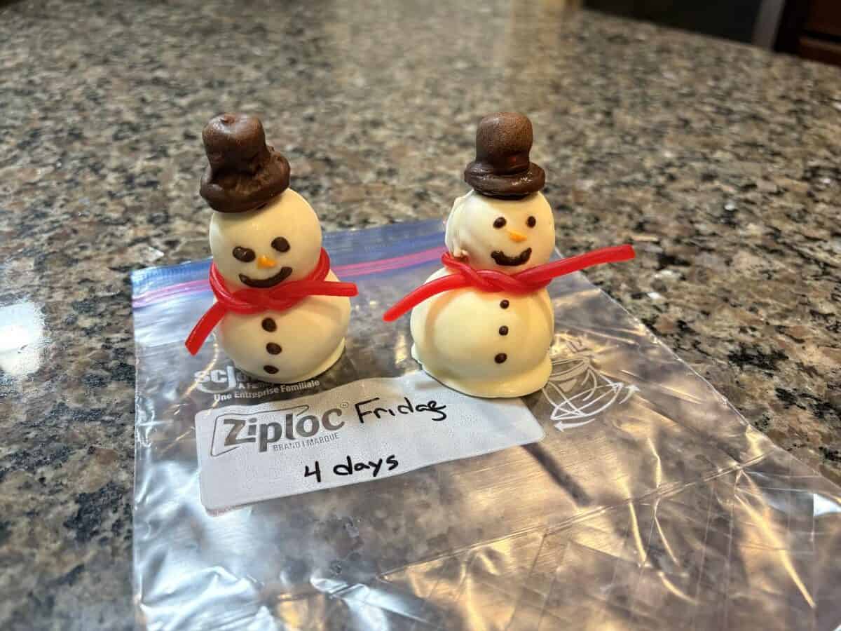 Two small Snowmen Truffles, each with a chocolate hat and a red licorice scarf, stand on a Ziploc bag labeled “Fridge 4 days” on a granite countertop.