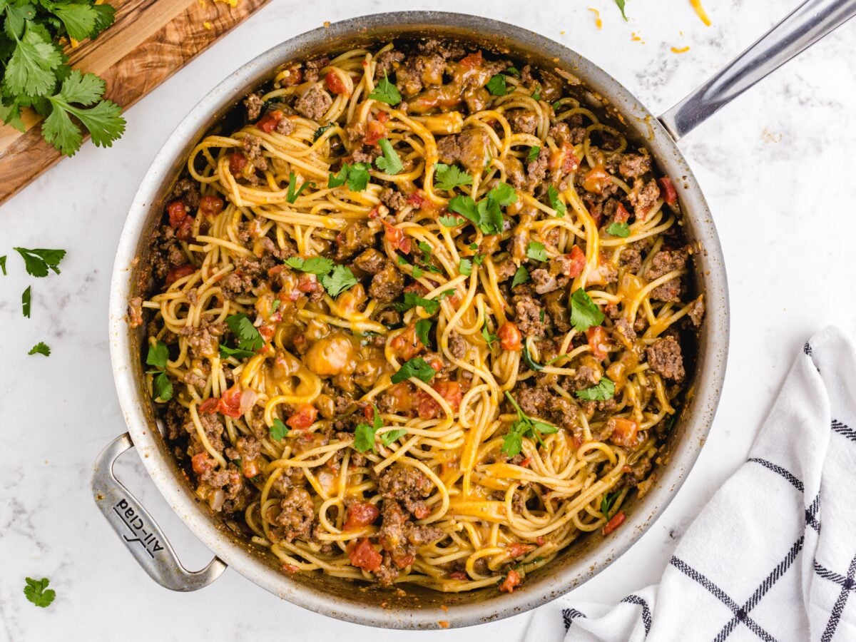 A skillet filled with Taco Spaghetti&mdash;cooked spaghetti mixed with ground beef, diced tomatoes, green peppers, melted cheese, and fresh parsley&mdash;sits on a white marble countertop next to a striped towel.