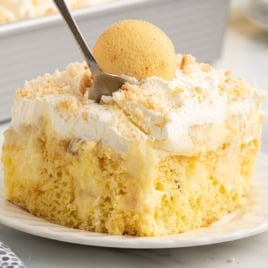 A slice of Banana Pudding Cake topped with whipped cream, crumbled cookies, and a whole vanilla wafer, with a fork taking a bite. The cake looks moist and creamy, served on a white plate—the perfect treat for anyone searching for the ultimate Banana Pudding Cake recipe.