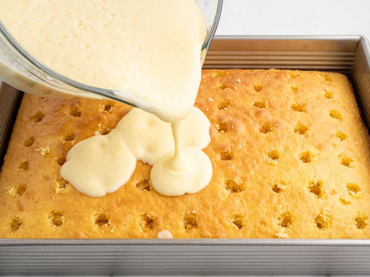 A glass bowl pours creamy filling over a yellow Banana Pudding Cake with evenly spaced holes in a square metal baking pan.