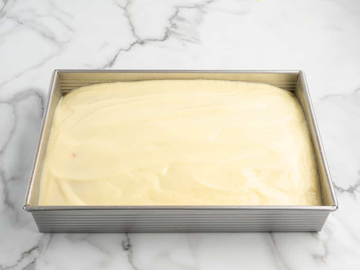 A rectangular metal baking pan filled with smooth, uncooked yellow cake batter for a delicious Banana Pudding Cake sits on a marble countertop.