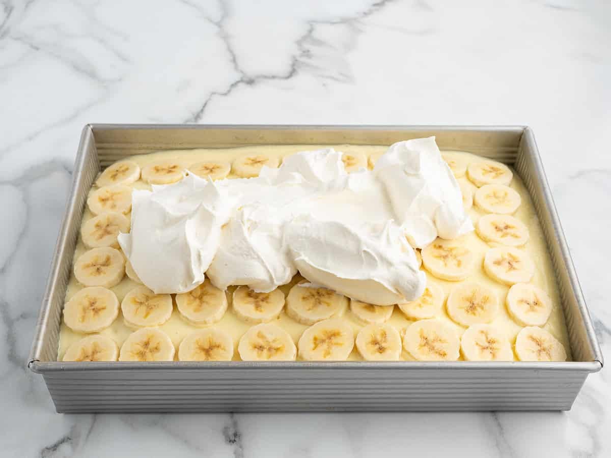 A rectangular pan filled with sliced bananas layered over creamy pudding, topped with a large dollop of whipped cream, sits on a marble countertop—an irresistible Banana Pudding Cake ready to be enjoyed.