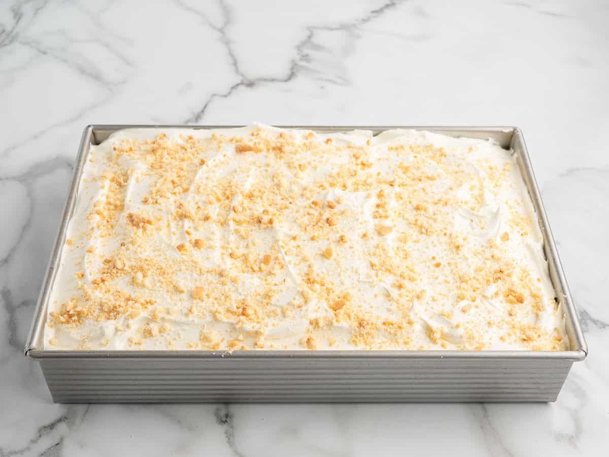A rectangular pan filled with a creamy Banana Pudding Cake, topped with crumbled cookies, sits on a white marble surface—an irresistible treat inspired by the classic Banana Pudding Cake Recipe.