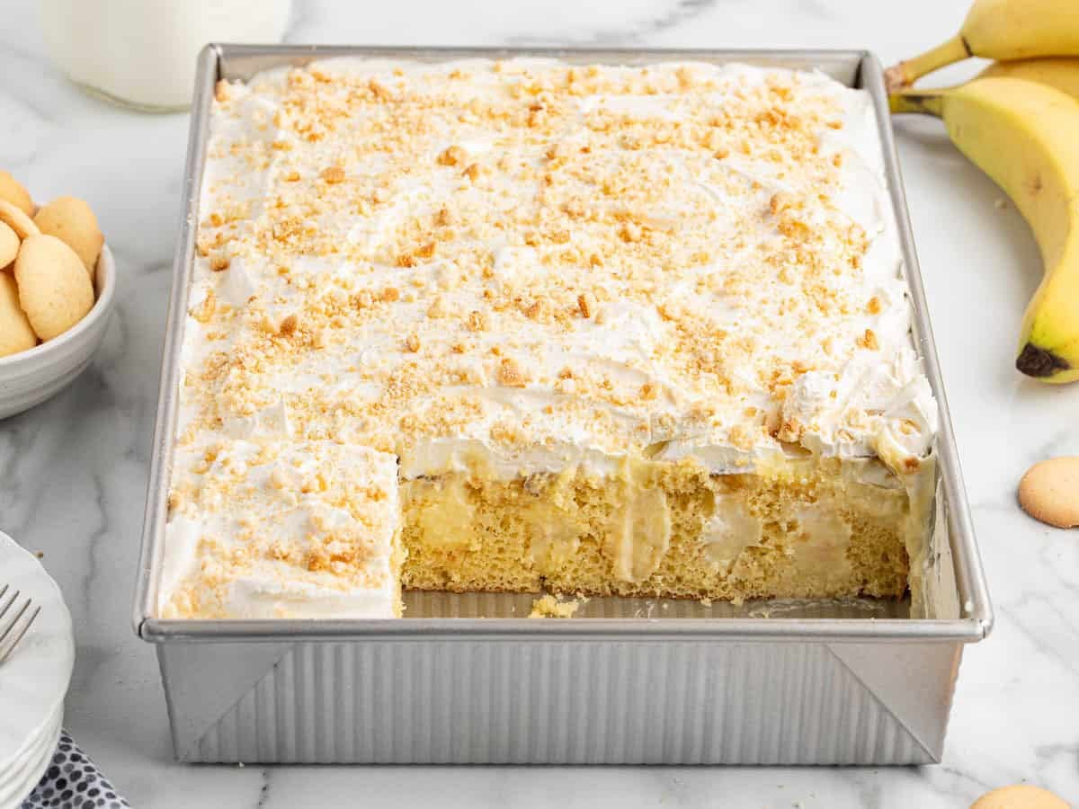 A square Banana Pudding Cake topped with whipped cream and cookie crumbs sits in a baking pan with one slice missing; bananas and cookies are nearby on a marble surface, making this Banana Pudding Cake Recipe irresistibly inviting.