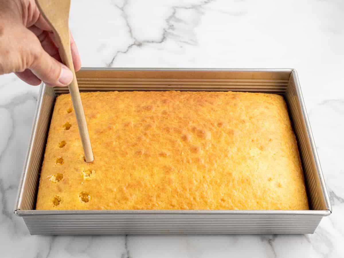 A hand uses a wooden spoon handle to poke holes in a rectangular yellow cake in a metal baking pan on a white marble surface, preparing it for the delicious Banana Pudding Cake Recipe.