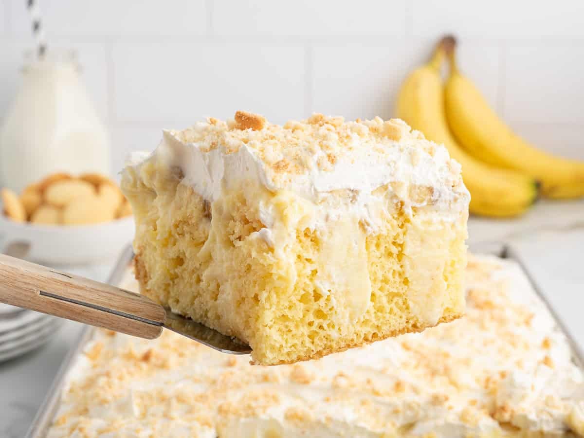 A close-up of a slice from our irresistible Banana Pudding Cake, topped with whipped topping and crushed cookies, is held above a sheet pan. Bananas and a bottle of milk in the background hint at the classic flavors in this beloved Banana Pudding Cake Recipe.