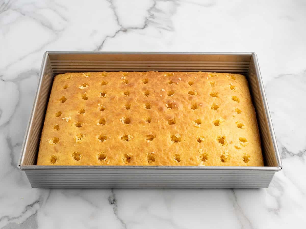 A Banana Pudding Cake, baked to perfection in a metal pan with evenly spaced holes poked into its surface, sits invitingly on a white marble countertop. This delectable treat is an irresistible take on the classic Banana Pudding Cake Recipe.