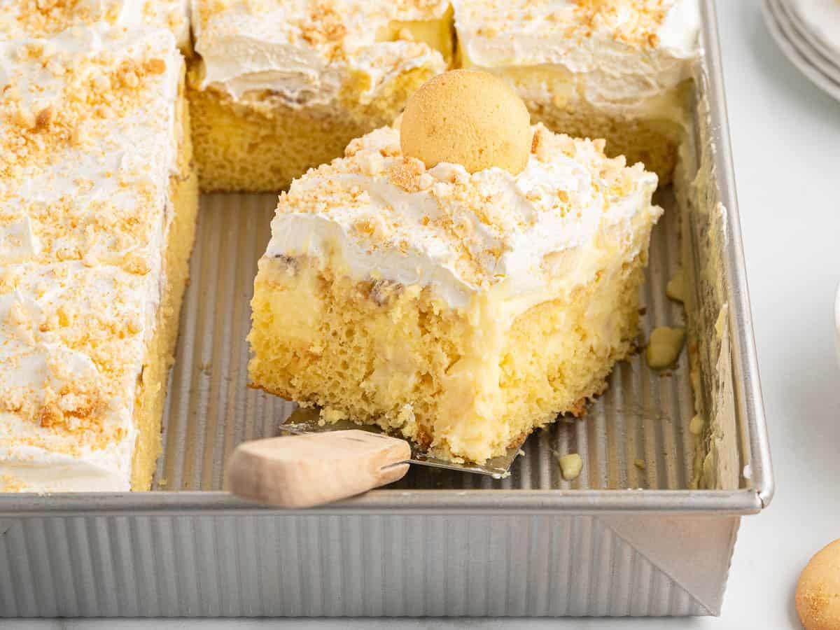 A square slice of Banana Pudding Cake, topped with whipped cream and a vanilla wafer, is lifted from a baking pan. Crushed cookies and creamy filling are visible, making this Banana Pudding Cake Recipe irresistibly delicious.